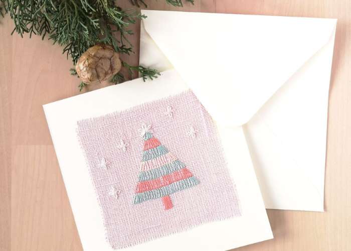 Carte postale avec broderie de sapin de Noël sur lin rose, enveloppe blanche et pomme de pin